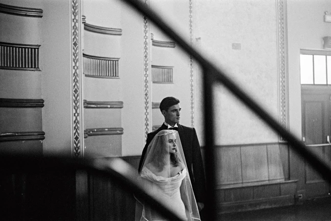 Black and white photograph of a bride and groom in front of paneled walls with decorative molding and window in a formal setting.