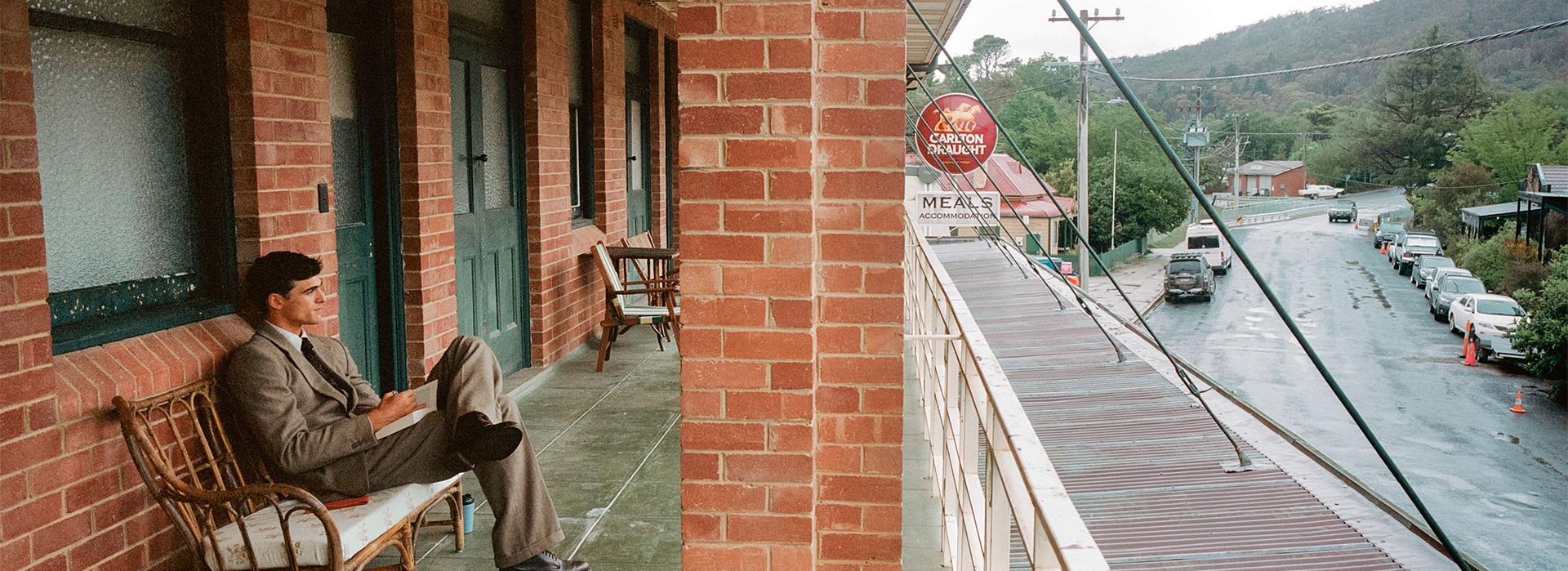 A man in a suit sits on a porch, reading. The backdrop is a street in a town with mountains in the distance and a vintage aesthetic.