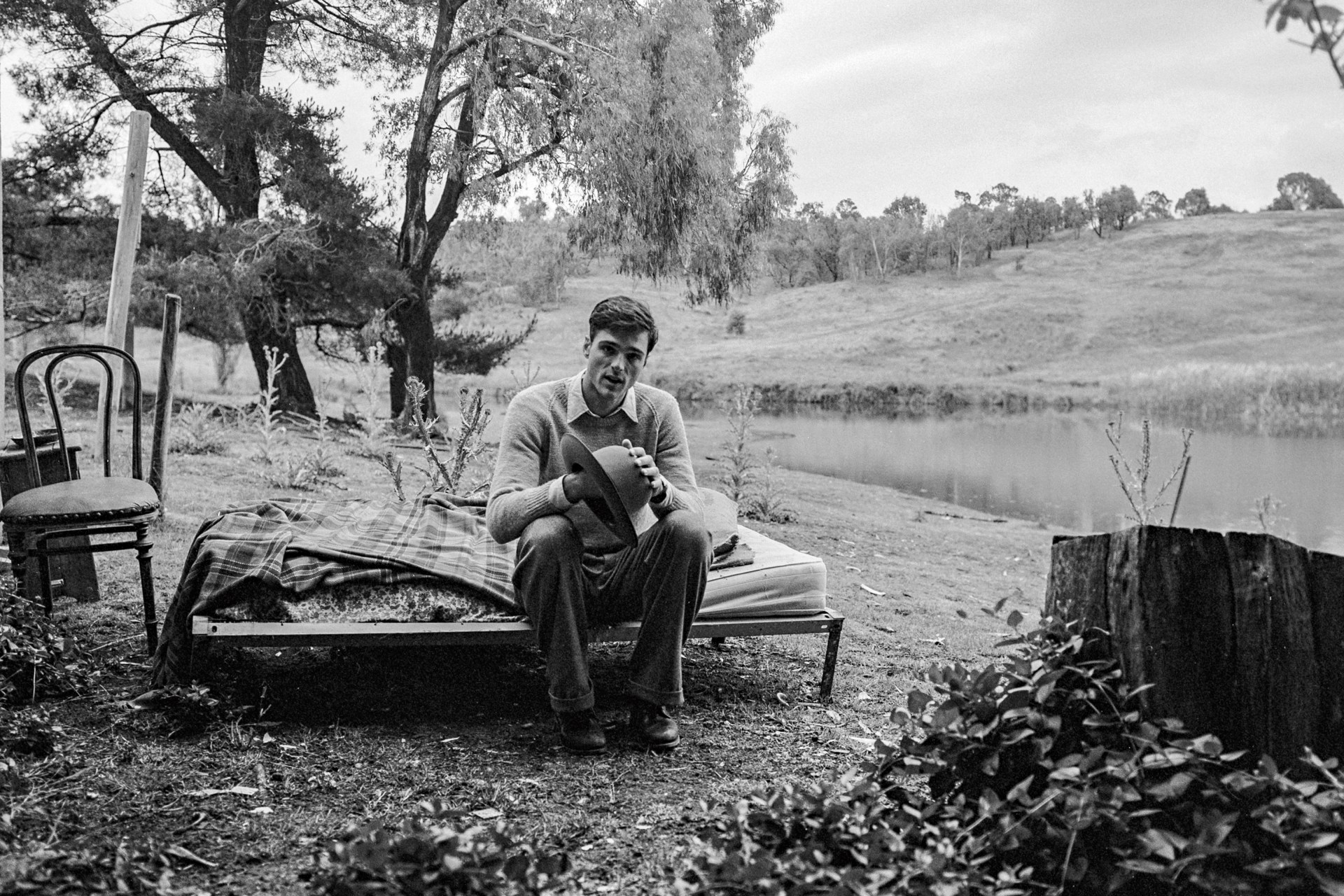 Monochrome portrait of a man sitting on an outdoor cot with a hat in his hands, by a lake, chair nearby, in a rural setting.