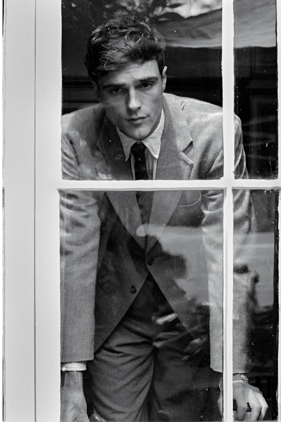 Jacob Elordi wearing a suit and tie looking through a multi-pane window in a black and white editorial portrait.