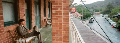 Jacob Elordi seated on a wicker bench on a brick building balcony overlooking a wet street.