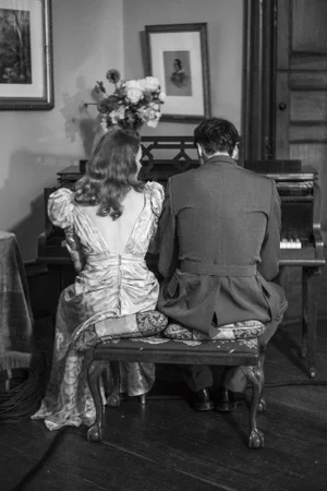 A man and woman seated at a piano together in a room, viewed from behind in a black and white photograph.