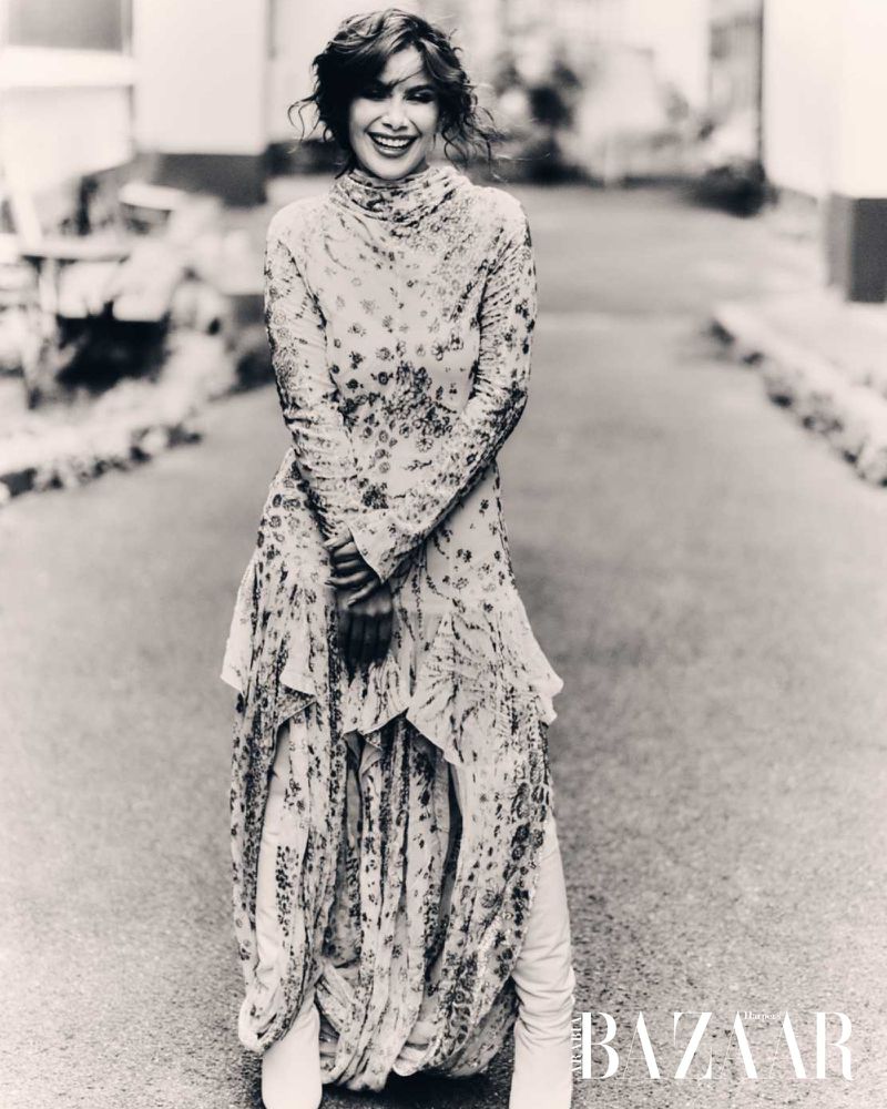 Actress Saba Mubarak smiling in a patterned turtleneck maxi dress and knee-high boots on an outdoor walkway in black and white.