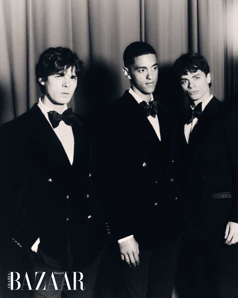 Three young men in formal wear, bow ties, and double-breasted jackets posing indoors against a curtained background in a black and white portrait.
