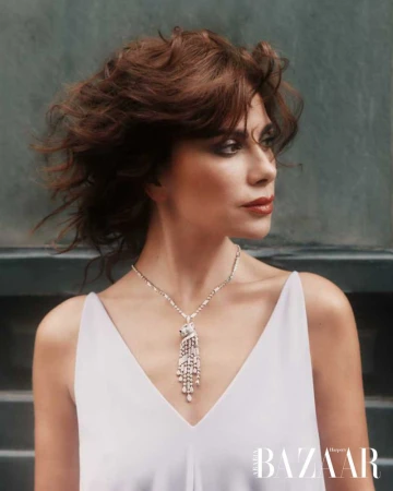 Close-up of a woman with auburn hair and red lipstick wearing a V-neck white dress and an ornate silver necklace in an indoor setting.