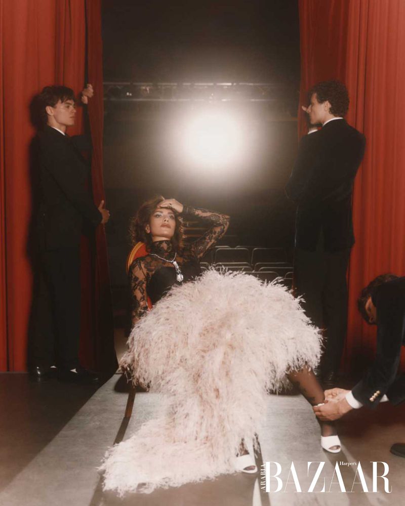 Woman in feathered dress seated on stage between two men opening red curtains, fourth man kneeling adjusting dress, illuminated by spotlight.
