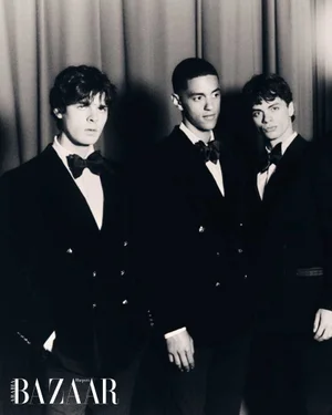 Three men in black formal tuxedos and bowties standing together in front of draped curtains in a black and white fashion portrait.