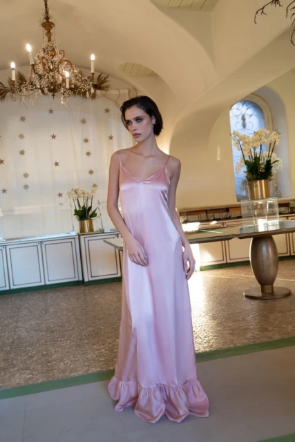 Elegant adult female in a long pink satin dress posing indoors near a window and a decorative table with orchids.