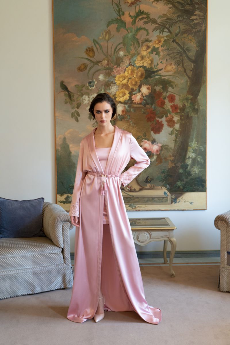 Adult woman poses in a pink satin robe and matching attire in an indoor setting with a floral painting on the wall. 