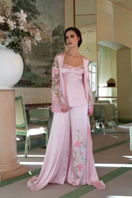 Elegant adult woman in a light pink satin lounge set, featuring floral embroidery, standing indoors with a classic interior design backdrop.