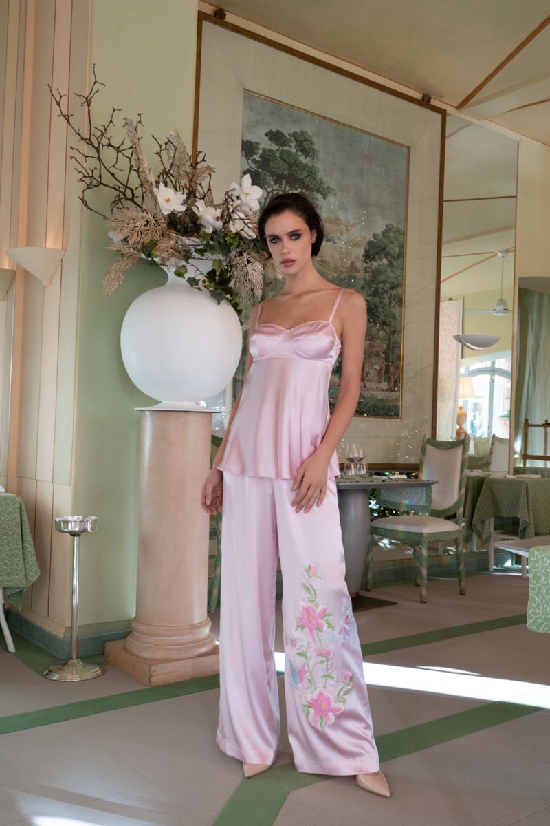 Model posing in a light pink satin camisole and matching wide-leg embroidered trousers in an elegant pastel interior.