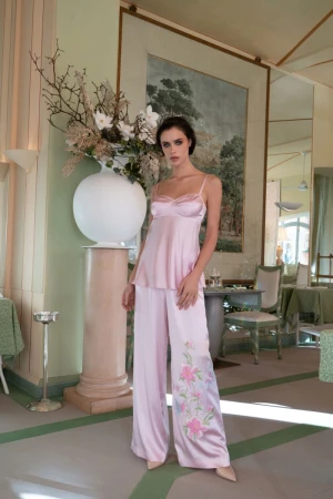 Model posing in a light pink satin camisole and matching wide-leg embroidered trousers in an elegant pastel interior.