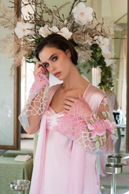 Elegant woman in a pink satin dress and floral-patterned lace sleeves poses in a room with a floral display and soft lighting.