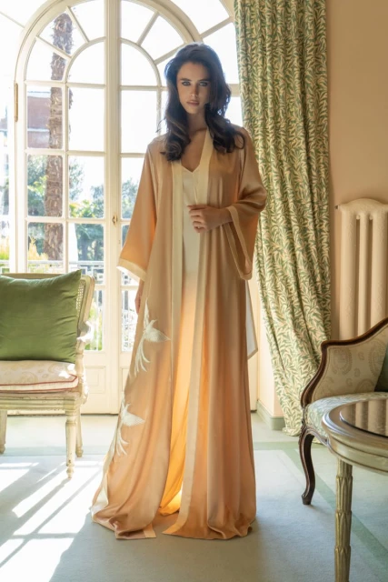 Elegant woman in peach-colored robe poses in a sunlit interior setting with decorative chair, curtains, and large window.