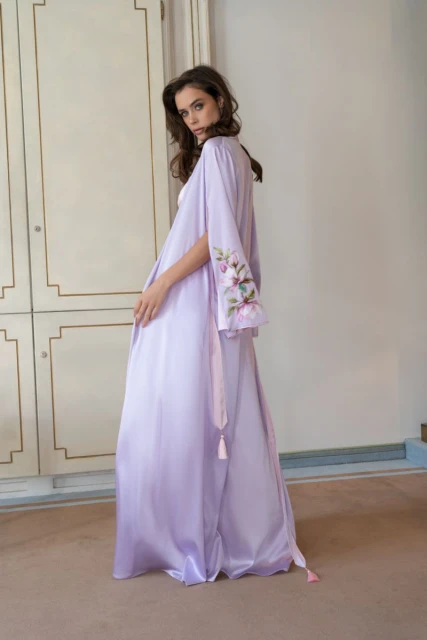 Elegant woman in a light purple robe with floral embroidery poses against a neutral-toned backdrop, showcasing fashion and style.