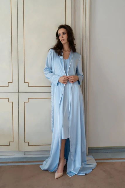 Elegant woman in a long blue robe stands indoors in front of paneled doors, wearing heels and gazing off to the side in a fashion pose.