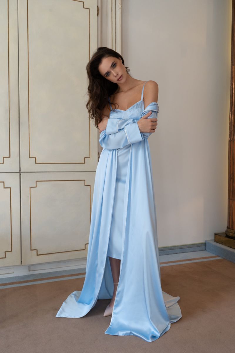 A brunette woman in a light blue satin gown and jacket, poses indoors in front of a light-colored wall with moulding details.