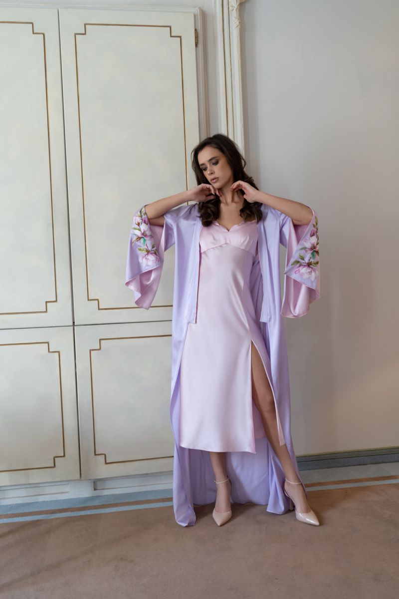 A woman poses in a lilac robe and dress indoors, hands framing her face, with light walls, light-colored shoes, and soft window light.