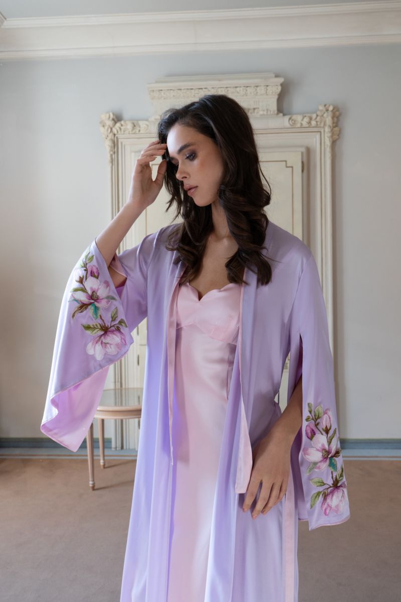 Woman in light pink slip dress and lavender robe with floral design in a neutral-toned interior setting, looking down with hand near her face.