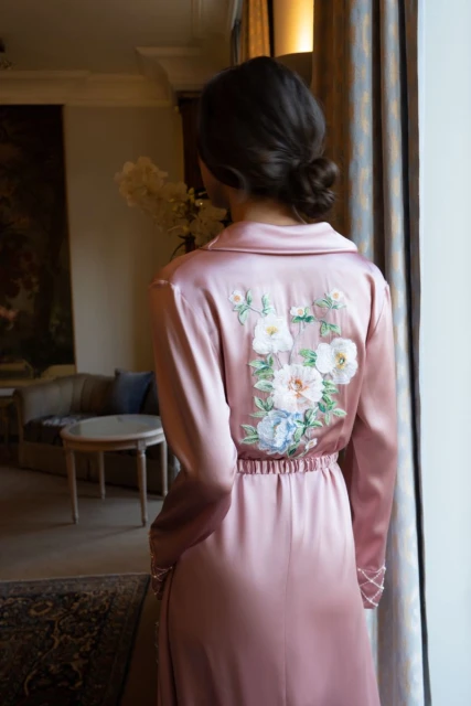 An adult female stands indoors, back to the camera, wearing a pink robe with floral embroidery. Light streams in from the window.