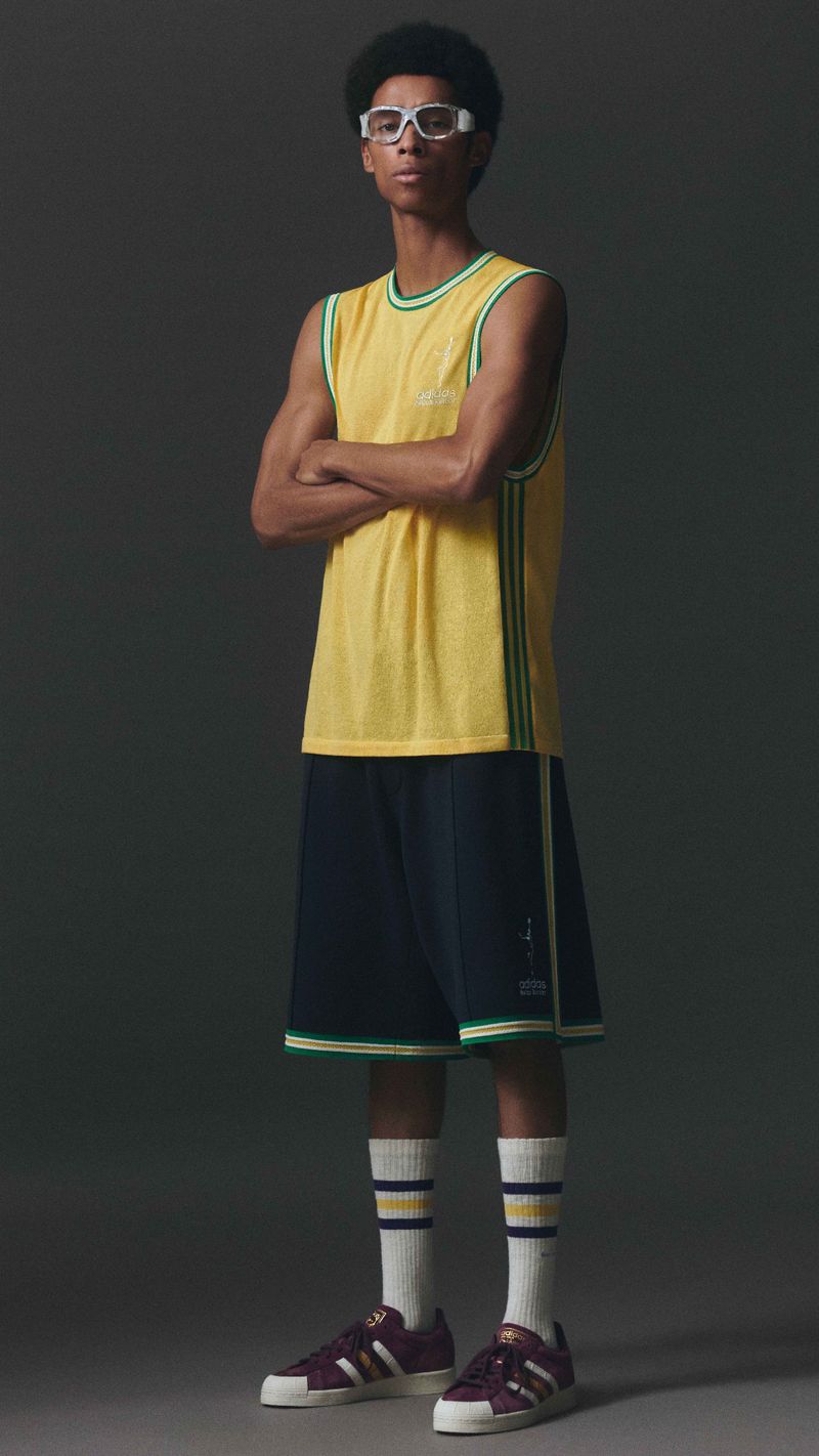 Full shot of a young adult male posing in a yellow jersey with blue shorts, striped socks and sneakers against a grey background.