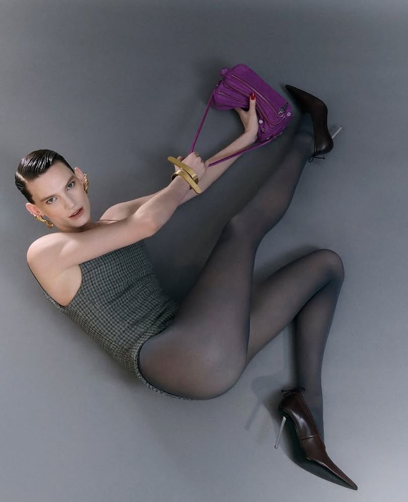 Fashionable person in a gray bodysuit, tights, and high heels, holding a purple purse while lying on a gray surface.