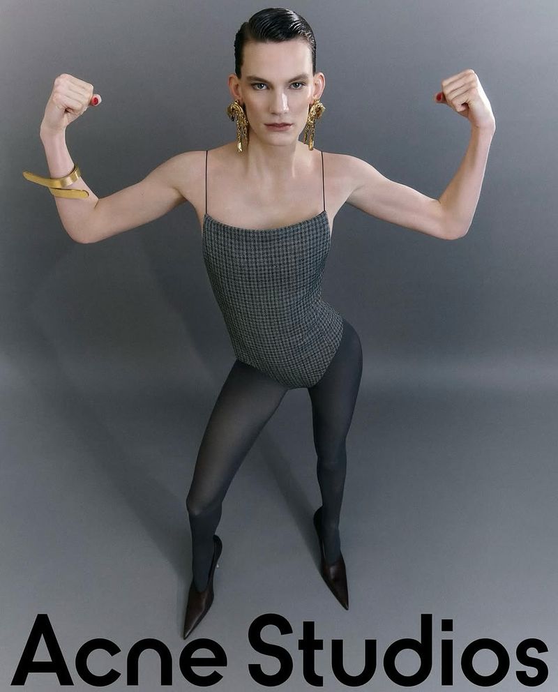 A fashion photo featuring a slender adult wearing a gray houndstooth leotard, dark tights, stiletto heels, and gold jewelry in a studio.