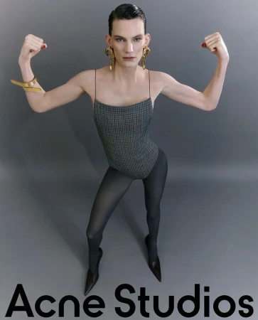 A fashion photo featuring a slender adult wearing a gray houndstooth leotard, dark tights, stiletto heels, and gold jewelry in a studio.