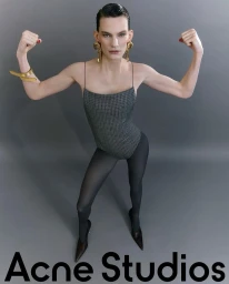 Lena Hardt posing with flexed arms in a grey bodysuit, black tights, pointed heels, and gold jewelry against a grey studio backdrop.