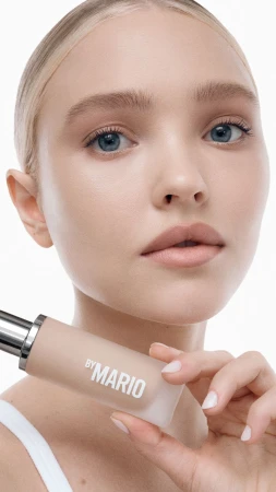 Blond woman holds By Mario foundation. Beauty shot emphasizes model's clean skin, product, and studio lighting against white backdrop.