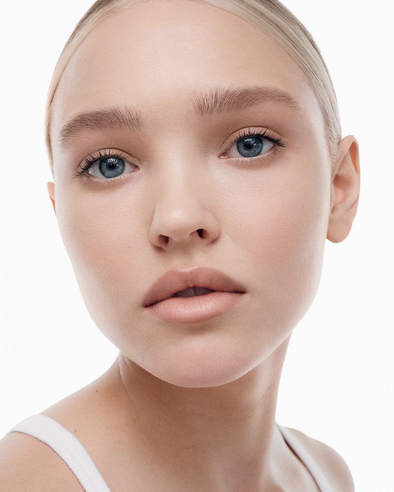 Close-up portrait of a fair-skinned young woman with blue eyes and blonde hair pulled back, showcasing soft, natural makeup against a white background.