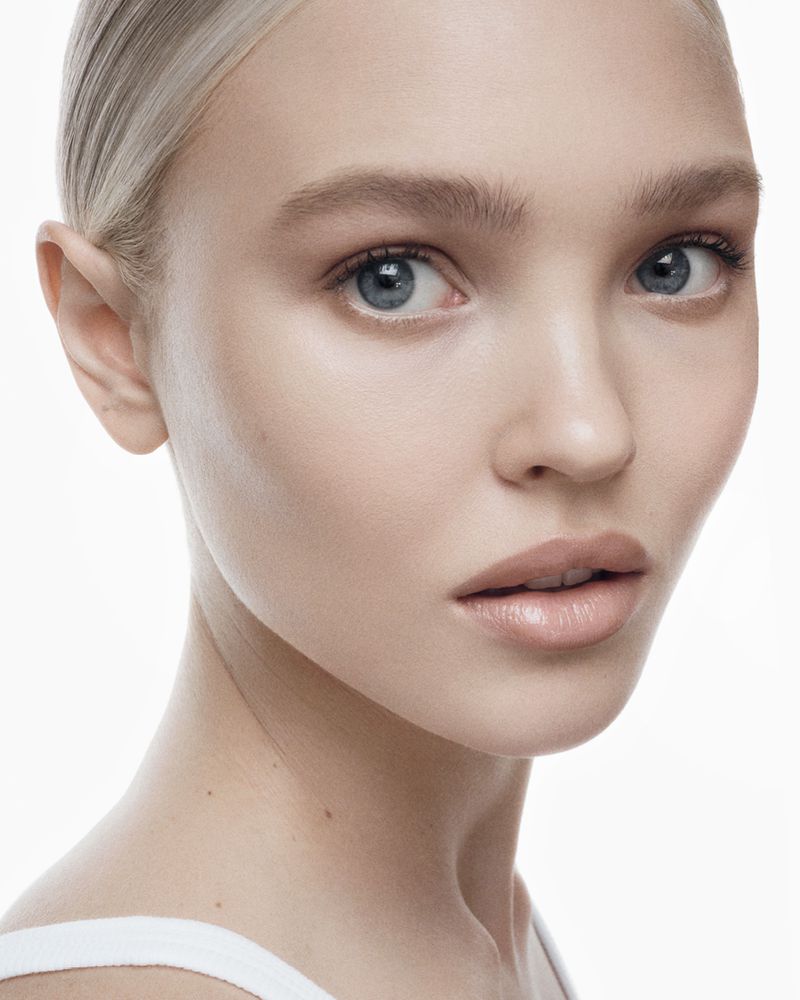Close-up portrait of an adult woman with blonde hair, blue eyes, and neutral expression against a bright white background in a beauty shot.