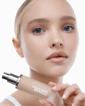 Close-up shot of a blonde-haired woman holding By Mario foundation, promoting beauty and makeup products against a white backdrop.