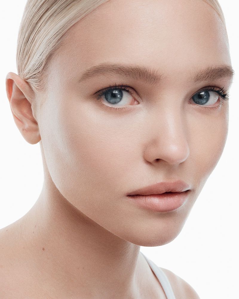 Close-up portrait of a blonde woman with blue eyes against a bright white background, focusing on skincare and beauty.