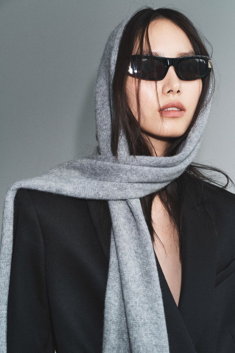 Portrait of an Asian woman with a gray scarf over her head, wearing sunglasses and a black blazer against a plain gray backdrop.