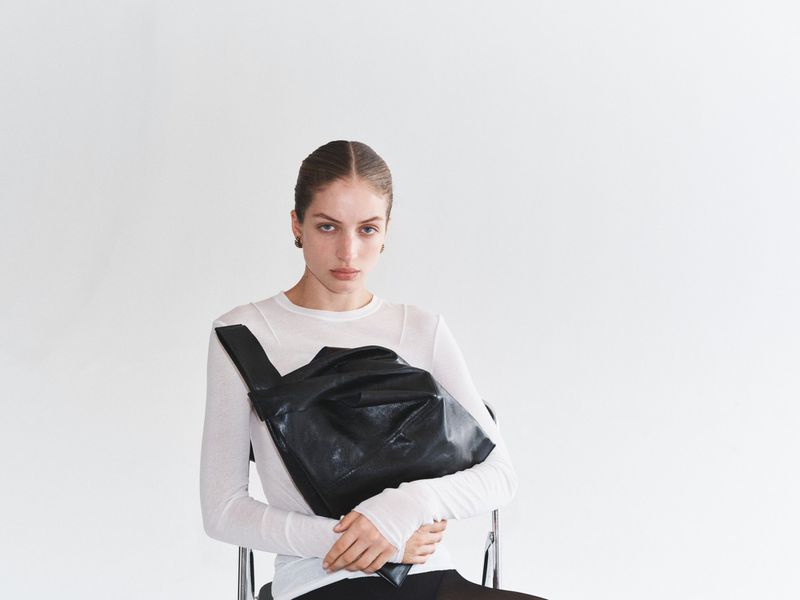 A woman with dark hair is seated, wearing a long-sleeved white shirt and black pants, holding a large black bag against a white background.