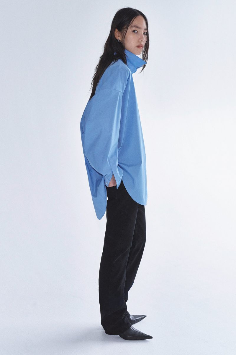 A full-length shot of an Asian woman in a blue top and black pants, posing in a photo studio against a white backdrop, looking at the camera.