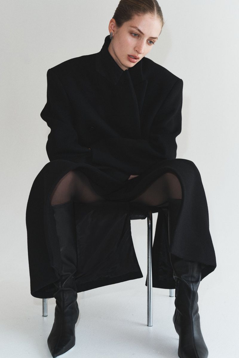A brunette woman with her hair pulled back is seated on a metal chair in a photo studio wearing a black coat and black boots.