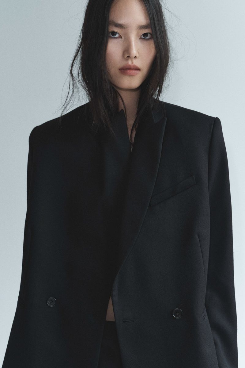Asian woman in oversized black blazer and dark top posing in a studio against a pale background. Fashion editorial style, portrait.