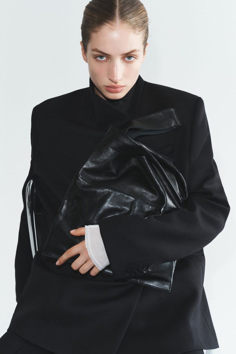 Portrait of a woman in a black blazer holding a shiny black bag in a bright, neutral setting, fashion editorial-style.