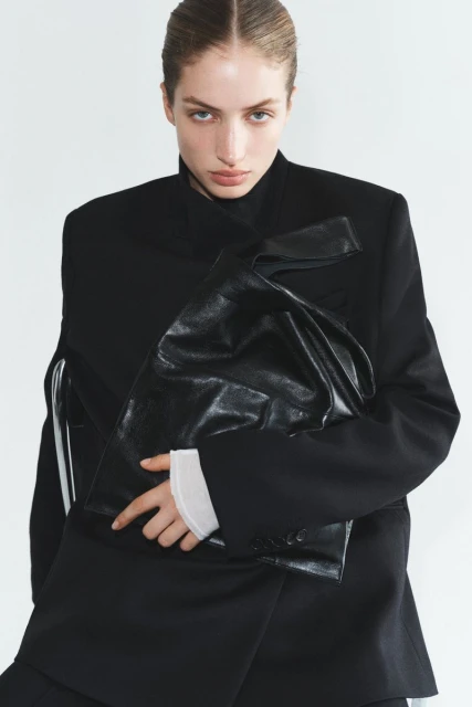 Portrait of a woman in a black blazer holding a shiny black bag in a bright, neutral setting, fashion editorial-style.