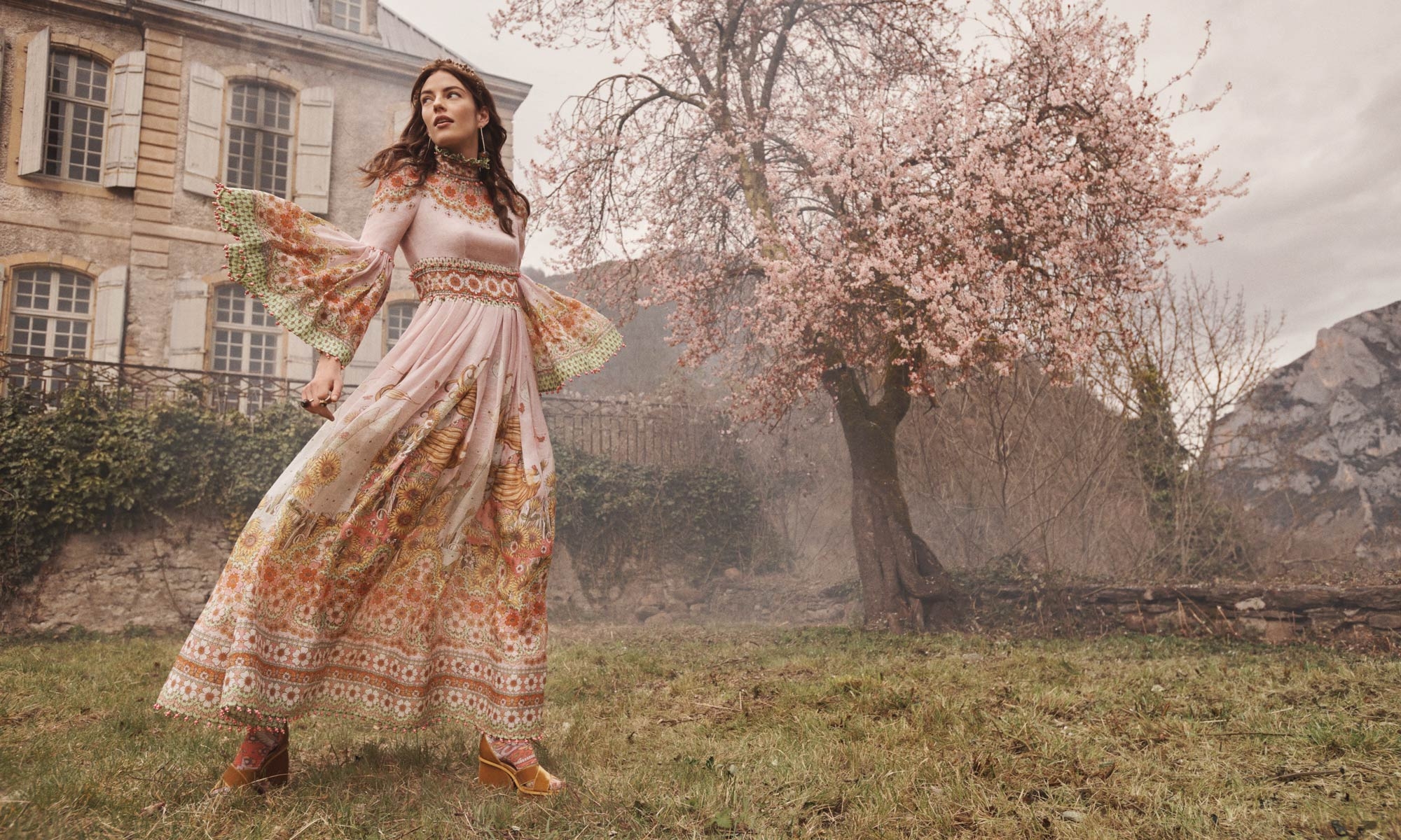 A woman in a floral dress stands in a field near a stone building and a blossoming tree on a cloudy day.
