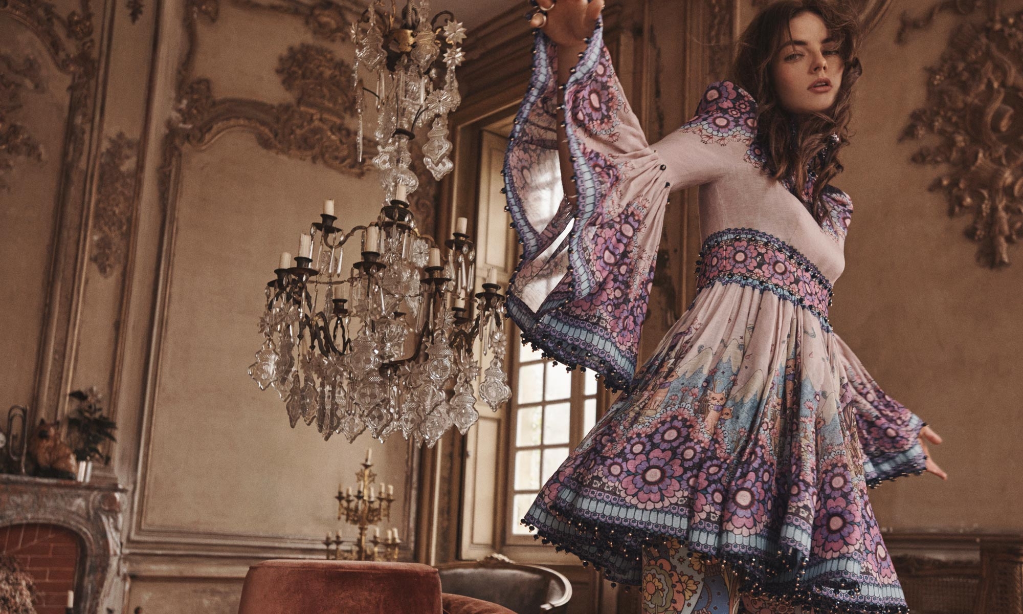 A woman poses indoors in a patterned bell-sleeve dress, with chandeliers and ornate wall details as a backdrop, in a fashion editorial shot.