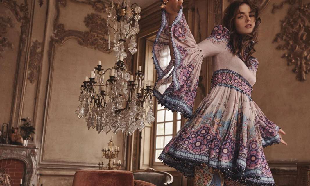 A woman poses indoors in a patterned bell-sleeve dress, with chandeliers and ornate wall details as a backdrop, in a fashion editorial shot.