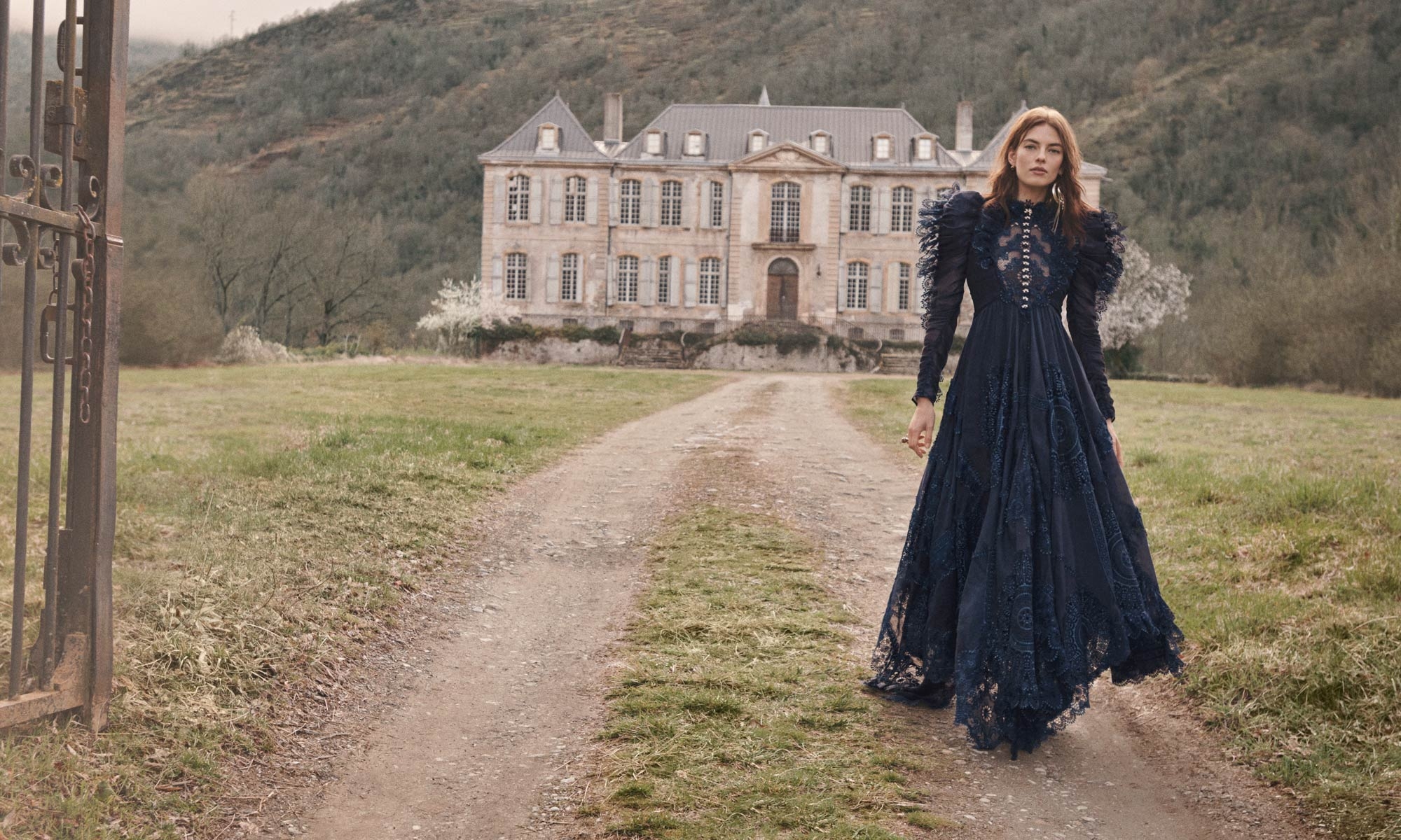 An adult female in a long dark dress walks on a dirt path toward a mansion, with a gate in the foreground and mountain in the distance.