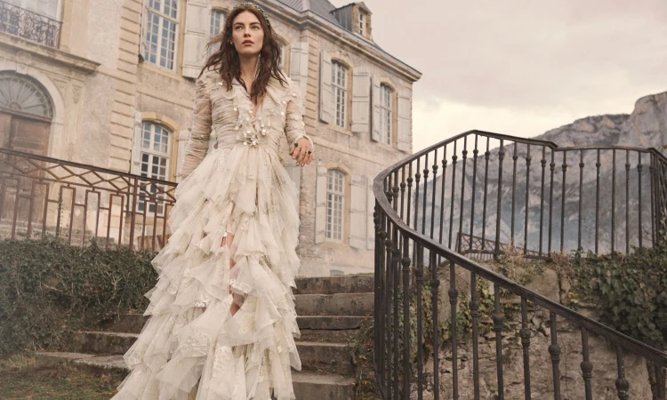 Model in ornate cream ruffled gown walking down stone steps before a large classical building.