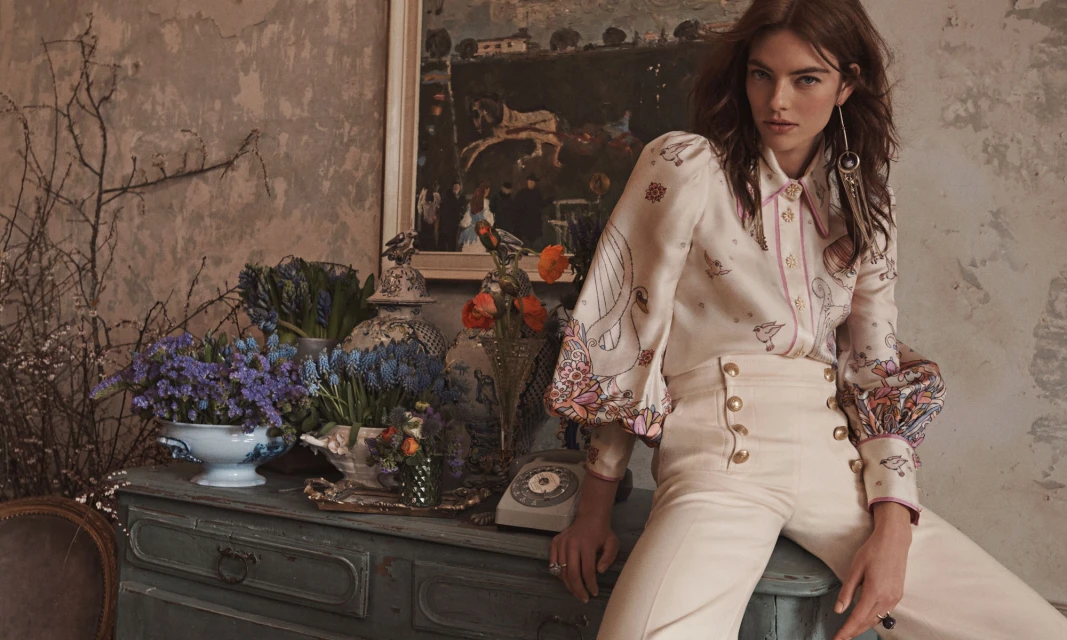 A model in a patterned blouse and cream-colored pants poses next to a blue credenza with a painting on the wall for a fashion editorial.