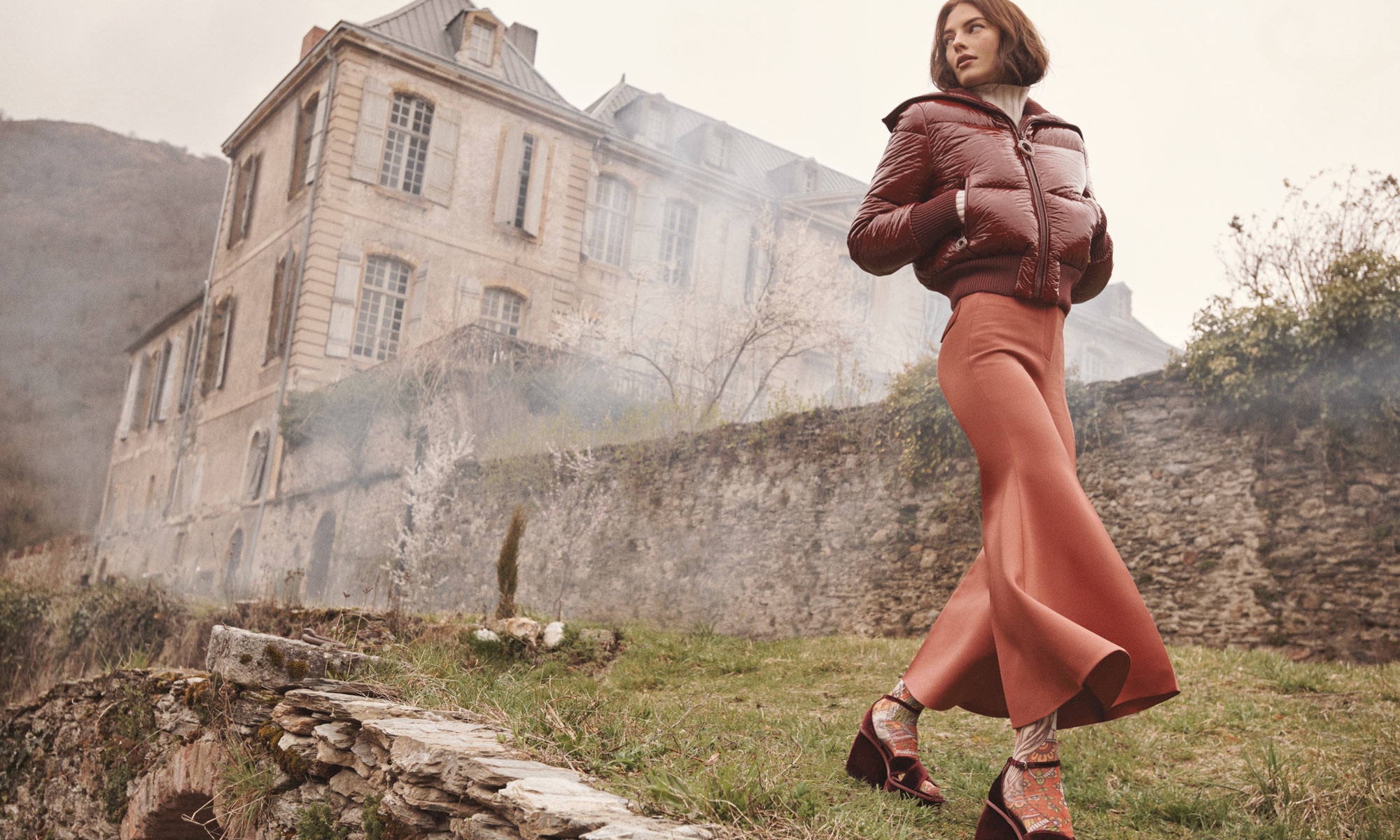 Model wearing a glossy dark red puffer jacket and terracotta skirt walking outdoors before a large stone building.