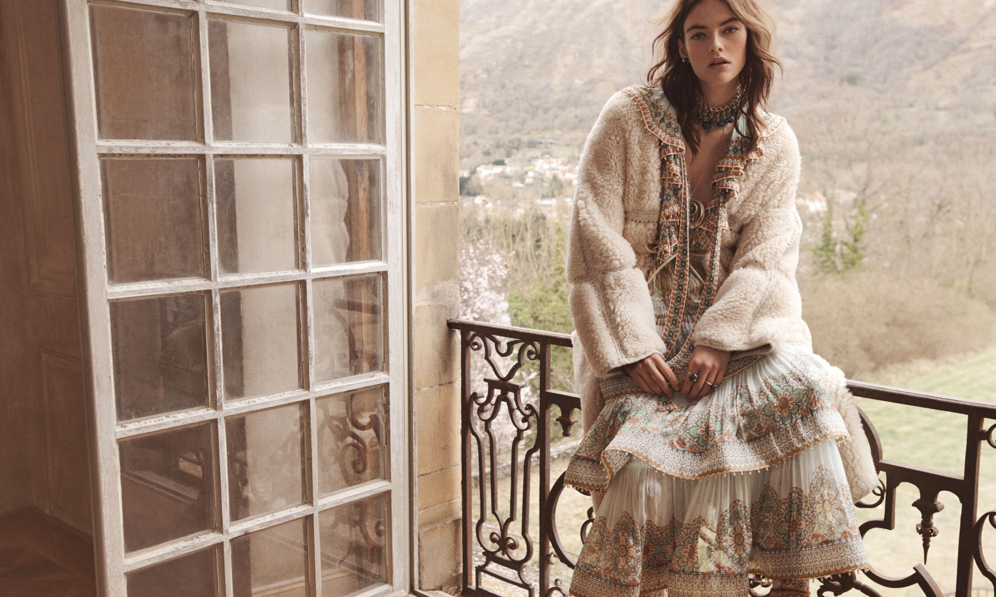 A fashionable woman poses on a balcony in a fuzzy coat and floral dress with a scenic view. The image has natural light and neutral colors.