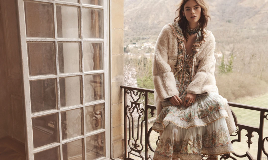 A fashionable woman poses on a balcony in a fuzzy coat and floral dress with a scenic view. The image has natural light and neutral colors.